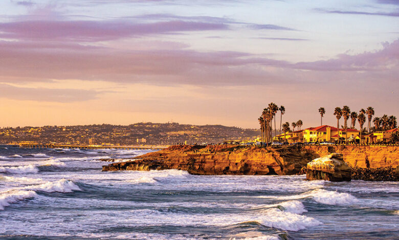 The San Diego coast seen from afar. There are cliffs with palm trees and blue sea with waves crashing. The sun is setting so there is a warm yellow glow