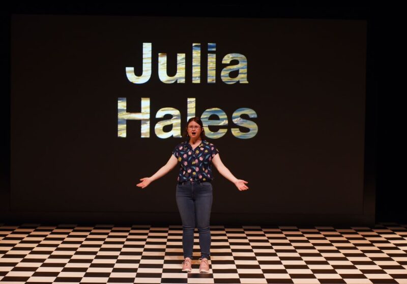 - Actress Julia Hales wearing glasses, blue jeans and a blue top with small flowers on a black and white chequer board stage in We Know We Belong Together with her name in silver letters on a black screen behind her