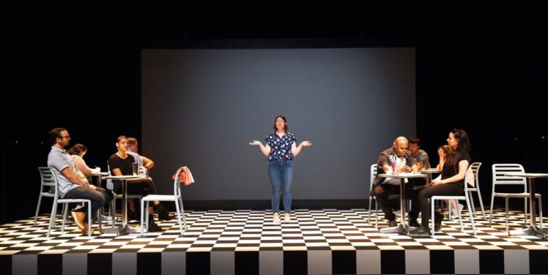 Cast of We Know We Belong Together, who all have Down's syndrome. on stage in a cafe setting with Julia Hales, the main character, in standing in the middle. There are four tables and two chairs at each. The background is black and there is a black and white chequered floor