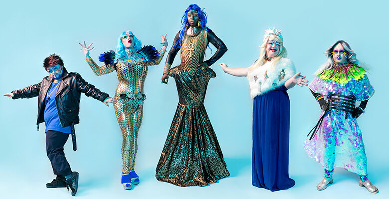 Five Drag Syndrome performers wearing glamourous, colourful and sparkly clothes, wigs and make up posing in front of a pale blue background