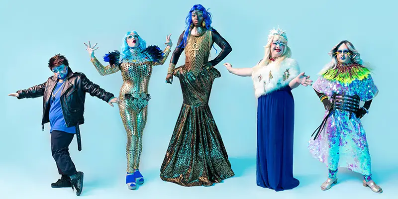 Five Drag Syndrome performers wearing glamourous, colourful and sparkly clothes, wigs and make up posing in front of a pale blue background
