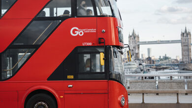 A red London bus viewed from the side driving across a bridge in London with the Tower of London in the background
