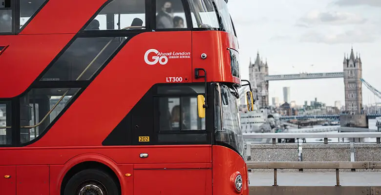 A red London bus viewed from the side driving across a bridge in London with the Tower of London in the background