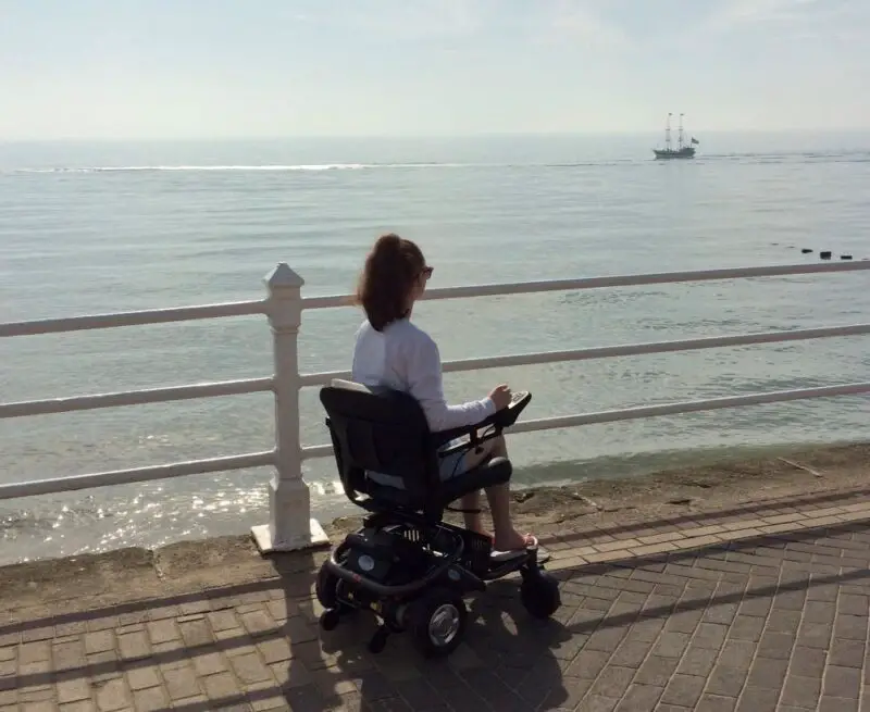 Pippa Stacey at Bridlington seafront sat in her powerchair looking out to sea. She is wearing a white, long sleeved shirt and her long, brown hair is tied in a ponytail