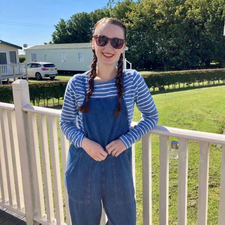 Pippa Stacey with long brown hair in two plats wearing blue dungarees, a blue and white striped top and sunglasses, smiling and standing in front of caravans in Bridlington