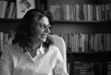 A black and whtie image of Shahd Alshammari with shoulder length hair wearing a white shirt and glasses looking to the side sat in front of a bookcase filled with books