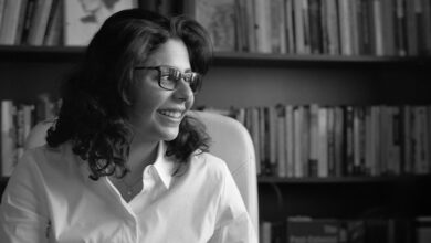 A black and whtie image of Shahd Alshammari with shoulder length hair wearing a white shirt and glasses looking to the side sat in front of a bookcase filled with books