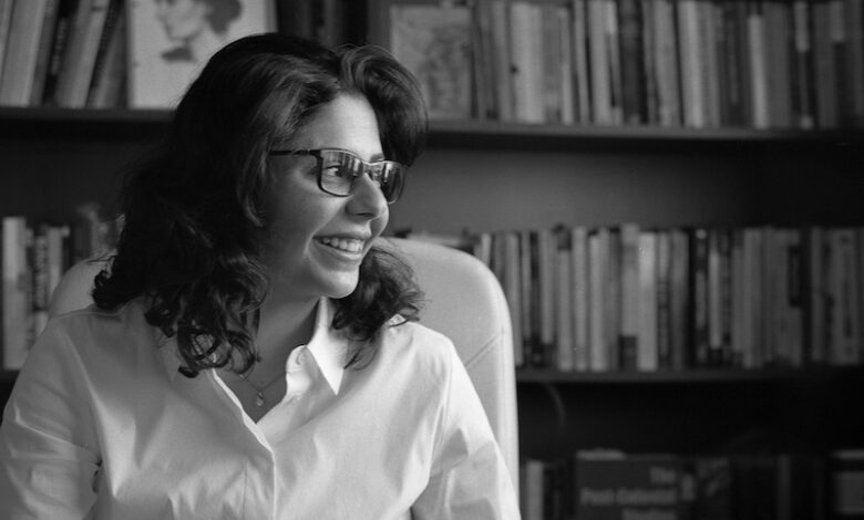 A black and whtie image of Shahd Alshammari with shoulder length hair wearing a white shirt and glasses looking to the side sat in front of a bookcase filled with books