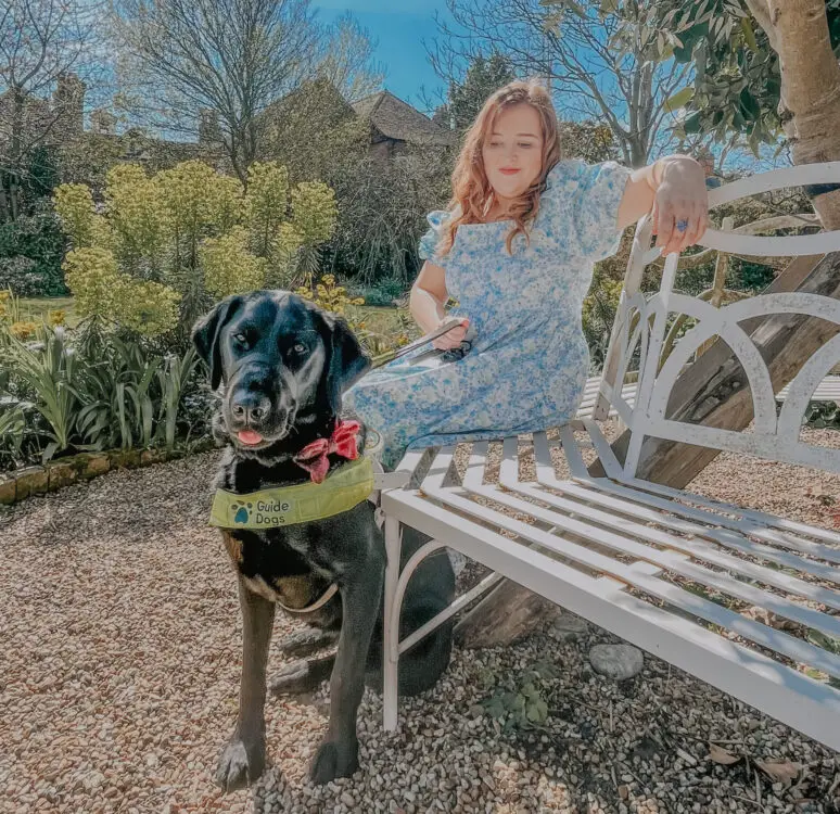Visually impaired blogger Emily Davison with her black guide dog sat on a white mental bench in a garden wearing a floral blue dress and with her long red hair down