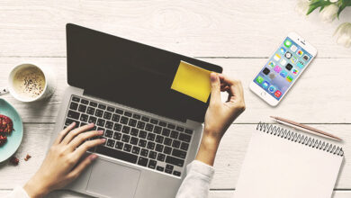 A silver laptop on a white desk viewed from above with a woman typing. There is a cup of coffee next, plate of cookies, phone and notepad to it