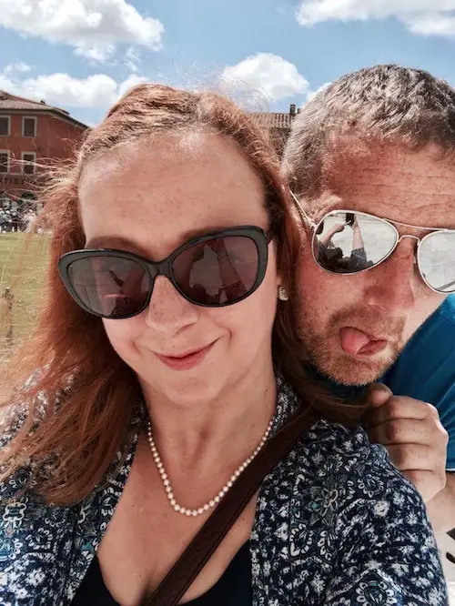 Carrie-Ann with her husband Darren behind her. They are both outside in the sun wearing sunglasses. Carrie-Ann is wearing a blue dress with a small white floral print and a pearl necklace and Darren is wearing a blue T-shirt