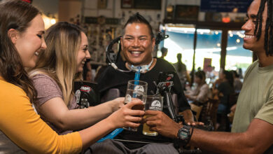 A man in a wheelchair with black hair, a black T-shirt and a tracheostomy with three friends around him all in a pub smiling and cheering with their drinks
