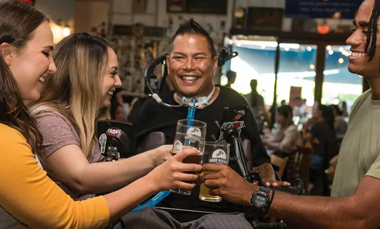 A man in a wheelchair with black hair, a black T-shirt and a tracheostomy with three friends around him all in a pub smiling and cheering with their drinks