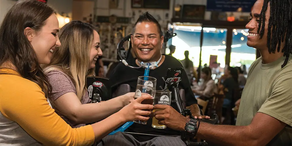 A man in a wheelchair with black hair, a black T-shirt and a tracheostomy with three friends around him all in a pub smiling and cheering with their drinks