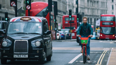 A busy road in London with a man on a bicycle, a black taxi and red busses and cars behind them
