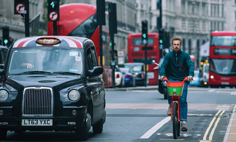 A busy road in London with a man on a bicycle, a black taxi and red busses and cars behind them