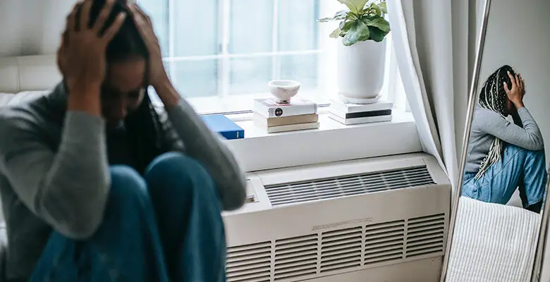 A woman sat on her bed with her head in her hands and her knees up to her chest. She is sat next to a heater below a window and window sill with books and a plant on it. She can be seen in a mirror on the other side of the room