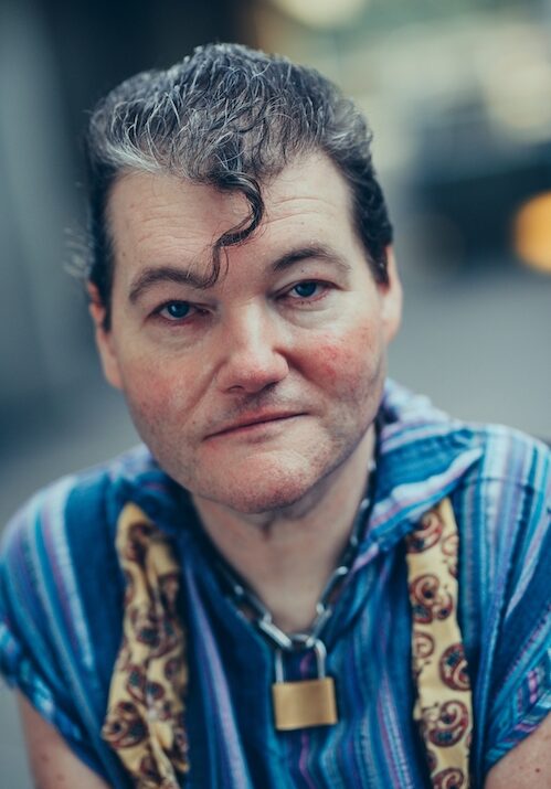 A head and shoulders photograph of John Kelly, white man, dark hair, signature kiss curl dropped onto forehead. Wearing a striped blue shirt with sleeves cut, chain and padlock necklace and a yellow scarf. John gazes past the camera