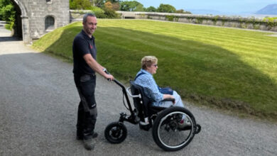 A woman sat in a Mountain Trike ePush wheelchair with a man stood behind the chair. They are on a gravel path with a grass verge in front of them, a stone wall and the sea below.