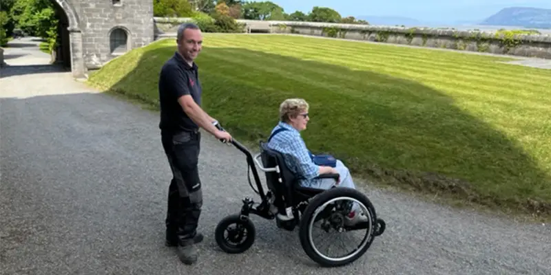 A woman sat in a Mountain Trike ePush wheelchair with a man stood behind the chair. They are on a gravel path with a grass verge in front of them, a stone wall and the sea below.