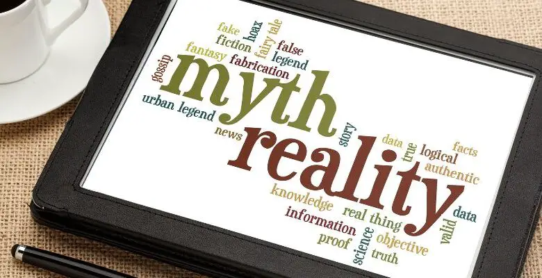 Myths and reality written in green and red on a tablet lying on a table next to a white cup of coffee and a stylus. There are other words around myth and reality, including logic, fiction, information, proof and false