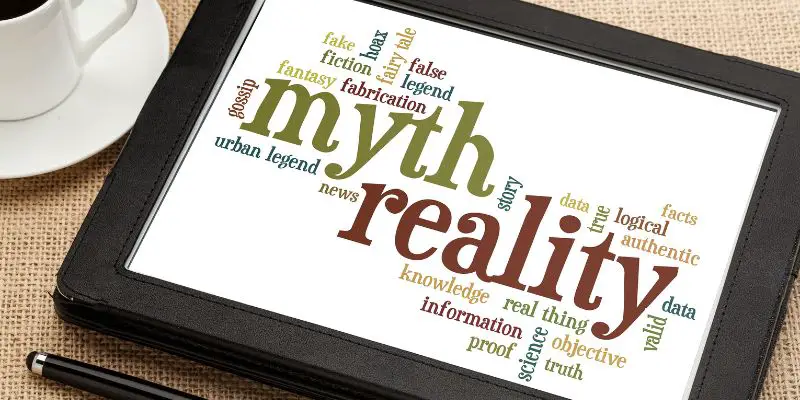 Myths and reality written in green and red on a tablet lying on a table next to a white cup of coffee and a stylus. There are other words around myth and reality, including logic, fiction, information, proof and false