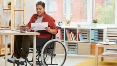 A woman in a wheelchair with short black hair wearing an orange shirt and black trousers sat at a desk in an office going through paperwork