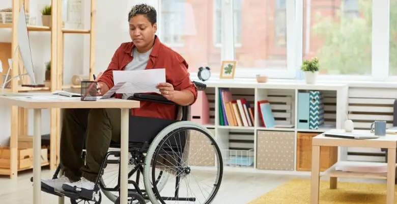 A woman in a wheelchair with short black hair wearing an orange shirt and black trousers sat at a desk in an office going through paperwork