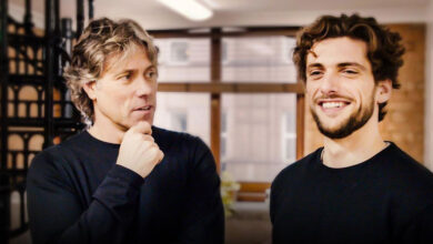 John Bishop with deaf son Joe. They both have dark curly hair and are wearing black jumpers. Joe is smiling and has a beard.