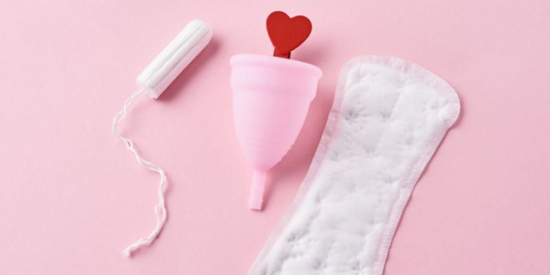A tampon, menstural cup with a red heart in it and a sanitary towel on a pale pink background