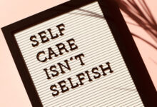 A white sign in a wooden frame that says 'Self care isn't selfish'. It's on a pink background with the shadow of a plant