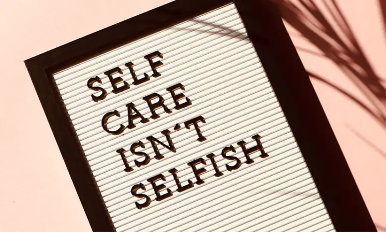 A white sign in a wooden frame that says 'Self care isn't selfish'. It's on a pink background with the shadow of a plant