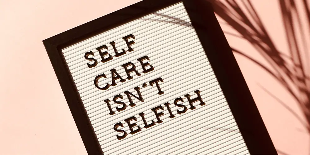 A white sign in a wooden frame that says 'Self care isn't selfish'. It's on a pink background with the shadow of a plant