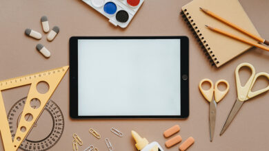 A collection of stationary and craft materials scattered across a table including a tablet, rubbers, glue, scissors, pencils, a notepad and paint palette