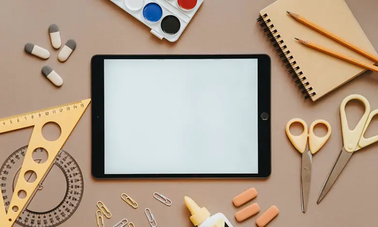 A collection of stationary and craft materials scattered across a table including a tablet, rubbers, glue, scissors, pencils, a notepad and paint palette