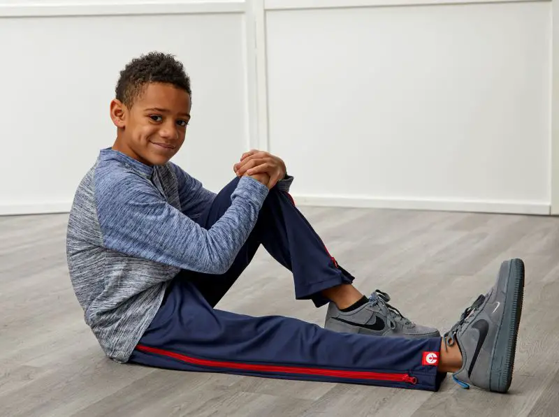 A young boy sitting on the floor wearing navy blue ZipOns trousers with a red zip and a blue and grey top
