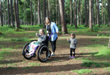 A mum with her two daughters walking in the woods. Her older daughter is in a Mountain Trike all-terrain wheelchair
