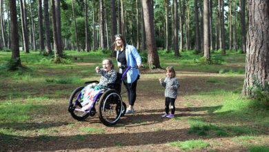 A mum with her two daughters walking in the woods. Her older daughter is in a Mountain Trike all-terrain wheelchair