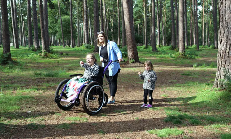 A mum with her two daughters walking in the woods. Her older daughter is in a Mountain Trike all-terrain wheelchair