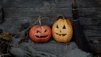 Two pumpkins with scary faces carved into them sat in front of a barn on an old grey blanket with a wooden broom next to them