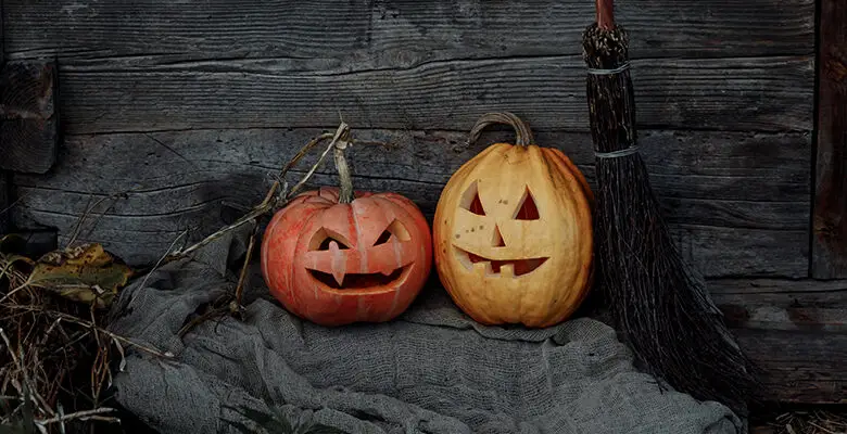 Two pumpkins with scary faces carved into them sat in front of a barn on an old grey blanket with a wooden broom next to them