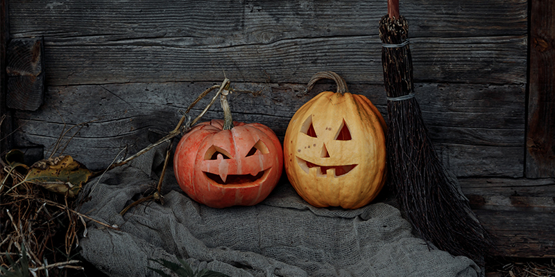 Two pumpkins with scary faces carved into them sat in front of a barn on an old grey blanket with a wooden broom next to them