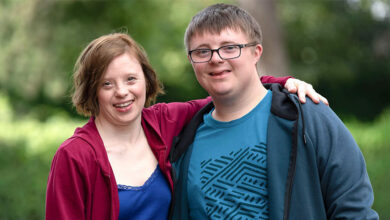 TV characters Ralph and Katie played by Leon Harrop and Sarah Gordy for a sitcom of the same name about marriage and Down's syndrome. Katie has short brown hair and is wearing a maroon cardigan and blue top and Ralph has short brown hair and is wearing black glasses, a turquoise top and blue cardigan