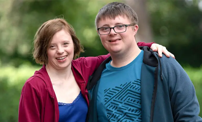 TV characters Ralph and Katie played by Leon Harrop and Sarah Gordy for a sitcom of the same name about marriage and Down's syndrome. Katie has short brown hair and is wearing a maroon cardigan and blue top and Ralph has short brown hair and is wearing black glasses, a turquoise top and blue cardigan