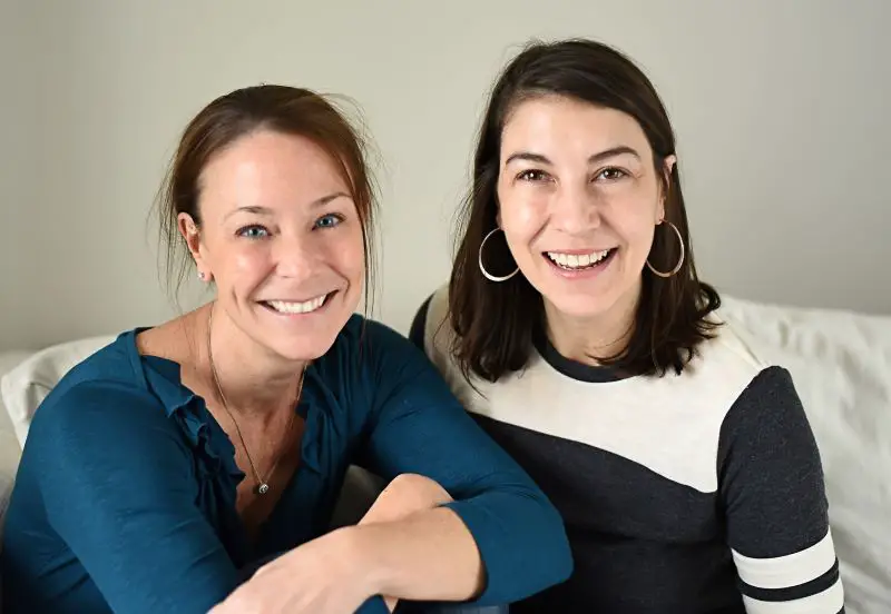 Nikki and Joanne, founders of BeFree adaptive clothing company, sat together on a sofa smiling at the camera. Nikki has her dark brown hair up and is wearing a petrol blue top with frills around the neck. Joanne has her shoulder-length black hair down, has large hoop earrings and is wearing a black and white top