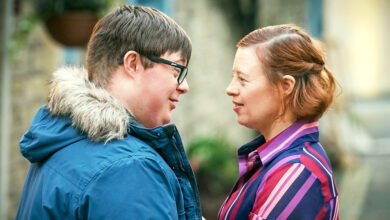 TV characters Ralph and Katie played by Leon Harrop and Sarah Gordy for a sitcom of the same name about marriage and Down's syndrome. Ralph and Katie are stood outside facing each other. Ralph has short brown hair, black glasses and a blue coat with fur-lined hood. Katie has shoulder-length red hair and is wearing a pink and red striped blouse