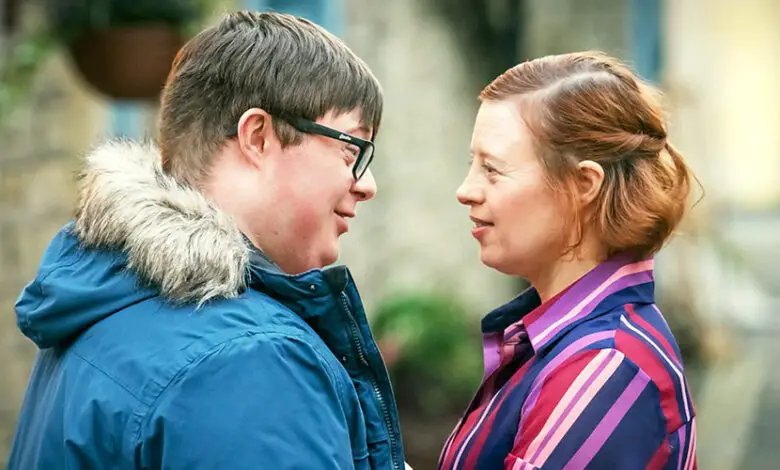 TV characters Ralph and Katie played by Leon Harrop and Sarah Gordy for a sitcom of the same name about marriage and Down's syndrome. Ralph and Katie are stood outside facing each other. Ralph has short brown hair, black glasses and a blue coat with fur-lined hood. Katie has shoulder-length red hair and is wearing a pink and red striped blouse