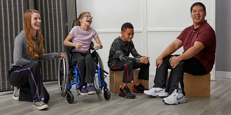 Four people - two children (a boy and girl) and two adults (a man and women) - wearing BeFree ZipOn adaptive trousers sitting and kneeling smiling at the camera