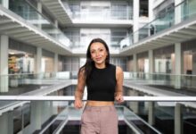 Natalie is a women of mixed heritage. She is standing inside at the Channel 4 offices with her elbows on the banister going across. Natalie is wearing a black crop top with beige trousers