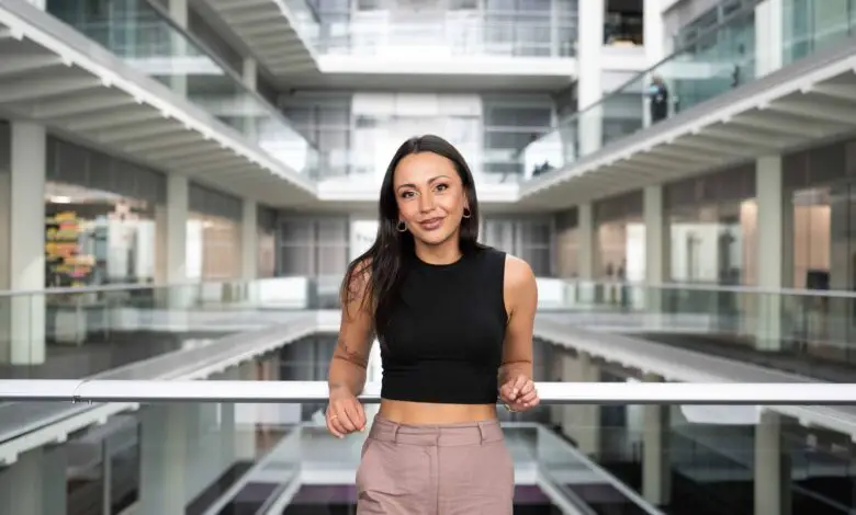 Natalie is a women of mixed heritage. She is standing inside at the Channel 4 offices with her elbows on the banister going across. Natalie is wearing a black crop top with beige trousers